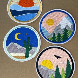 Op de afbeelding: Vier geborduurde patches met natuurscènes. Eén patch toont een zonsondergang boven water, een andere een berglandschap met een zon, een derde een woestijnnachtscène en de laatste een zonsondergang met bomen en een vogel.