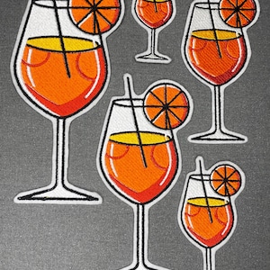 Op de afbeelding: Vijf geborduurde patches met een cocktail in een glas op een voet met een feloranje vloeistof, een gele bovenlaag, een zwart rietje en een schijfje sinaasappel als garnering. De patches variëren in grootte op een donkergrijze achtergrond.