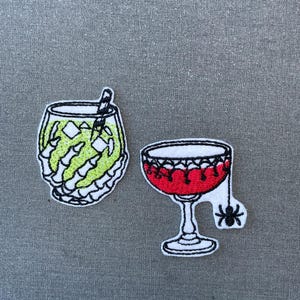 Può includere: Due toppe ricamate a tema cocktail. Una toppa mostra un bicchiere pieno di liquido verde lime tenuto da una mano scheletrica bianca con una cannuccia a righe. La seconda toppa raffigura una bevanda rossa in un calice con un ragno nero.
