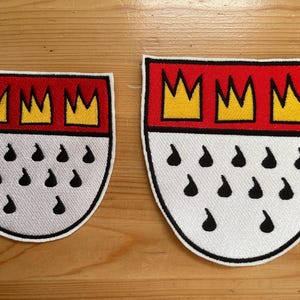以下が含まれることがあります： ドイツ、ケルンの紋章が描かれた2つの刺繍パッチ。各パッチは、3つの黄色の王冠が付いた赤い上部、黒い涙の形をした白い下部、黒い縁取りで構成されています。