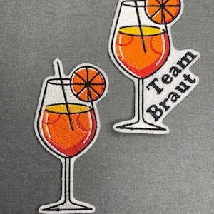 May include: Two embroidered patches depicting cocktail glasses with orange slices. The glasses are filled with an orange liquid and have a straw. One patch has the text "Team Braut" in black.