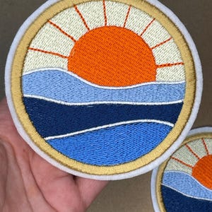 Op de afbeelding: Geborduurde patch met een zonsondergang boven oceaangolven. Het ontwerp omvat een oranje zon met stralen, blauwe en marinegolven en een gouden rand. De patch is rond met een witte rand.