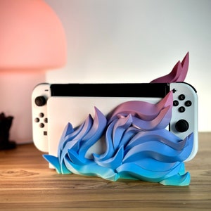 May include: A blue and pink 3D printed stand for a Nintendo Switch console. The stand is shaped like a stylized flame with a gradient of colors. The Switch console is sitting in the stand.
