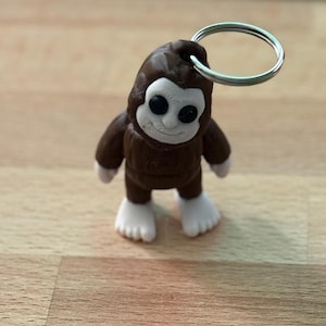 May include: A brown Bigfoot keychain with a silver key ring. The Bigfoot figure has a white face, white feet, and black eyes. The figure is standing on a wooden surface.