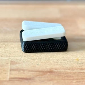 May include: A black and white 3D printed foot pedal with two white buttons. The pedal has a textured surface.