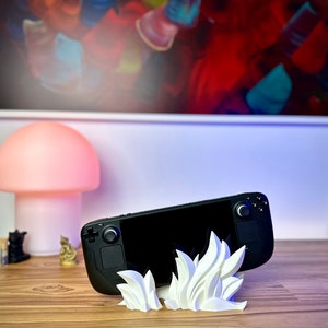 May include: A black handheld gaming console with white buttons and a black screen is propped up on a white 3D printed stand shaped like flames. The console is on a wooden surface.