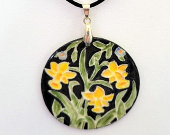 May include: A black cord necklace with a round pendant featuring a floral design. The pendant is painted with yellow daffodils, green leaves, and black and white accents on a black background.
