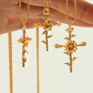 Könnte beinhalten: Drei goldene Halsketten mit floralen Kreuzanhängern. Die Kreuze sind mit kleinen Blumen und Blättern verziert. Die Ketten sind zart und haben ein geflochtenes Design.