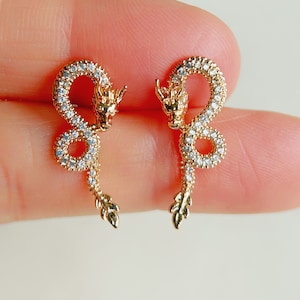 May include: A pair of gold-tone dragon earrings with clear crystal accents. The earrings are in the shape of a dragon with its head facing forward and its tail curled around its body.