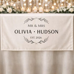May include: Cream-colored table runner with black text and a decorative wreath design. The text reads "MR & MRS OLIVIA • HUDSON EST. 2026". The runner is displayed on a table with candles and floral arrangements, suitable for weddings and events.