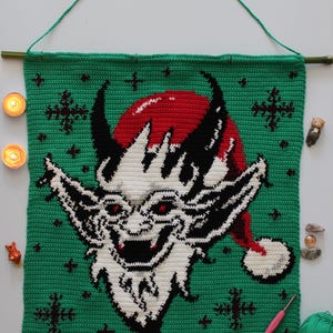 May include: Crocheted wall hanging featuring a devilish face with a red Santa hat on a green background. The artwork is adorned with black horns, white facial features, and red eyes. The wall hanging is suspended from a bamboo rod with a green cord.