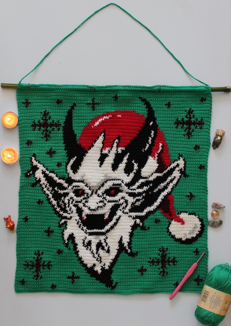 May include: Crocheted wall hanging featuring a devilish face with a red Santa hat on a green background. The artwork is adorned with black horns, white facial features, and red eyes. The wall hanging is suspended from a bamboo rod with a green cord.