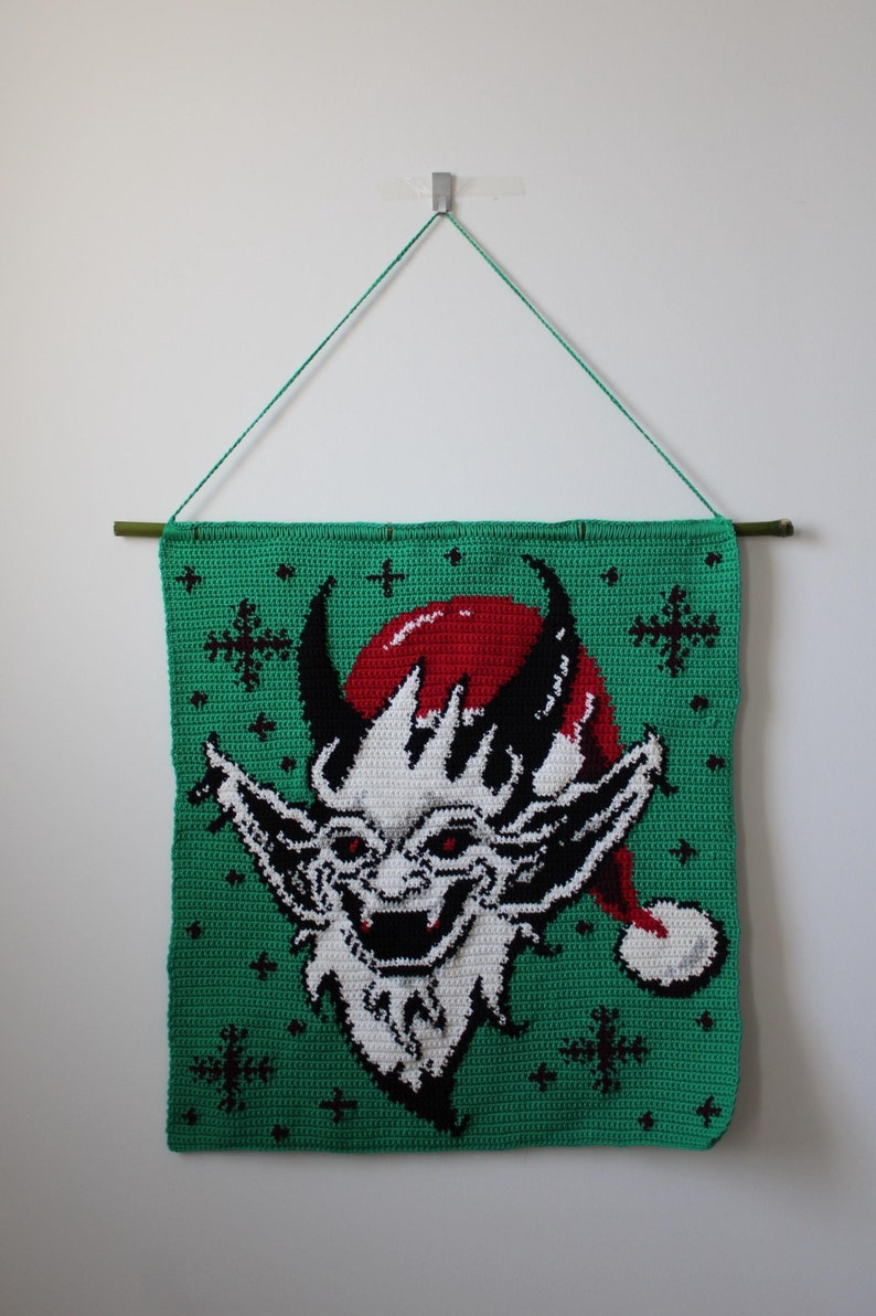 May include: A green crocheted wall hanging featuring a devilish face wearing a red Santa hat. The face is white with black details and red eyes. The background is green with black snowflake accents. A green cord is used for hanging.