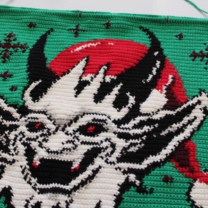 May include: Crocheted wall hanging featuring a devilish creature with a red Santa hat, black horns, and white and black facial details. The background is green with red and brown snowflake accents. A wooden dowel is at the top.