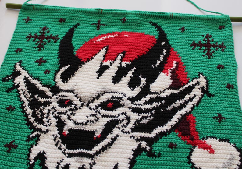 May include: Crocheted wall hanging featuring a devilish creature with a red Santa hat, black horns, and white and black facial details. The background is green with red and brown snowflake accents. A wooden dowel is at the top.