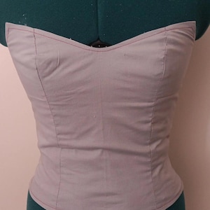 May include: A light gray corset top with a sweetheart neckline, displayed on a green dress form. The corset features red stitching along the seams, highlighting its tailored construction. The fabric appears to be a soft cotton or similar material.