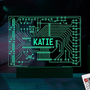 May include: A green circuit board design with the name "KATIE" in white letters. The circuit board is illuminated with a green light and is sitting on a wooden base. A remote control with multiple colored buttons is in the foreground.