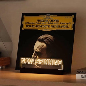 May include: A black vinyl record with a gold label featuring a portrait of Frederic Chopin and the text "FREDERIC CHOPIN 10 Mazurkas - Prélude op. 45 - Ballade op. 23 - Scherzo op. 31 ARTURO BENEDETTI MICHELANGELI STEREO" is displayed on a wooden surface with a neon sign that reads "NOW SPINNING".