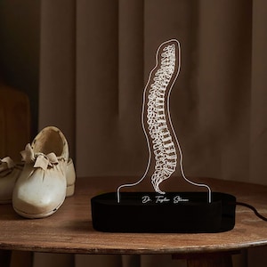 Puede incluir: Una lámpara negra impresa en 3D con forma de columna vertebral humana con el texto "Dr. Taylor Stone" en la base. La lámpara está sobre una mesa de madera con dos zapatos blancos en primer plano.