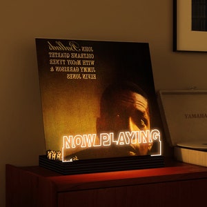 May include: A black illuminated vinyl record display with the words "NOW PLAYING" in white neon. The record features the album "Ballads" by John Coltrane Quartet. The display sits on a wooden surface, with a vintage record player in the background.