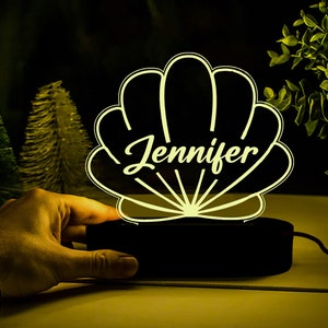 以下が含まれることがあります： 「Jennifer」という名前が刻まれた白い貝殻の形をしたナイトライト。ライトは黒いベースに置かれ、コンセントに差し込まれています。
