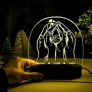 May include: A white illuminated night light with a design of two hands holding a baby. The base of the night light is black and has the names "David", "Caleb", and "Katie" inscribed on it.