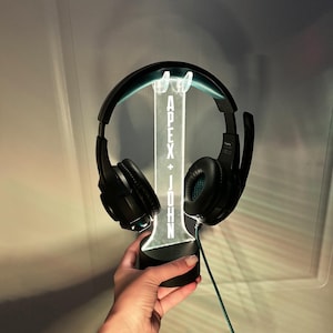 May include: A black gaming headset with a microphone is displayed on a black and white LED stand. The stand has the words "Apex + John" illuminated on it.