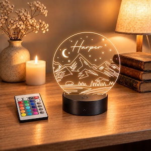 May include: A circular acrylic night light featuring an engraved mountain landscape, a crescent moon, stars, and a bear silhouette with the name Harper. The light sits on a black circular base and includes a multi-coloured remote control.