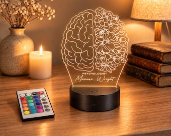Puede incluir: Una lámpara LED de acrílico con un diseño grabado de un cerebro humano a la izquierda y patrones florales a la derecha. El texto dice PSYCHOLOGIST Manasi Wright. La lámpara se asienta sobre una base circular negra e incluye un mando.