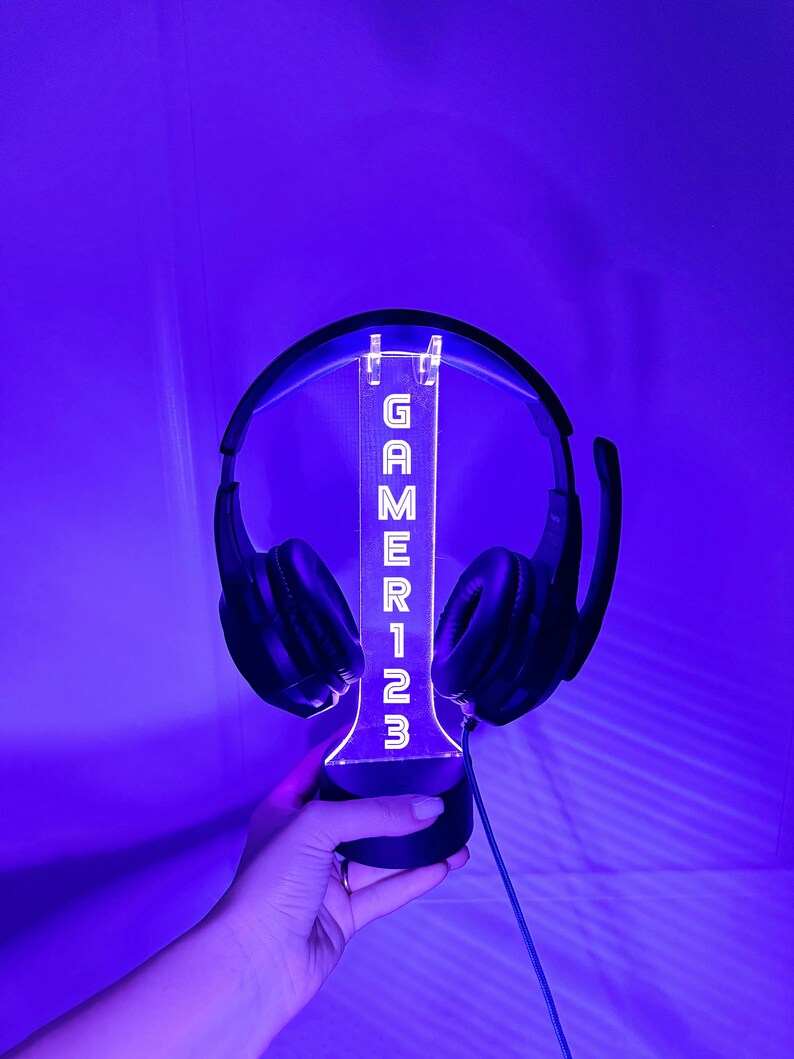 May include: A black headset with a clear acrylic stand that has the word "GAMER123" printed on it in white. The stand is lit up with a blue light.