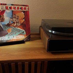 May include: A vinyl record album cover for "The Best of Kansas" is displayed on a wooden surface. The album art features a man with a long beard, a ship, and the words "NOW PLAYING". A vintage turntable is to the right.