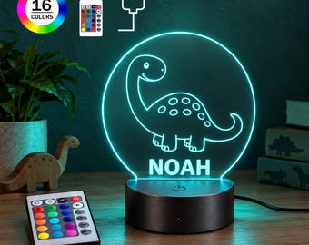 Puede incluir: Una luz nocturna LED circular con el contorno grabado de un dinosaurio y el nombre NOAH en letras mayúsculas. La lámpara se asienta sobre una base cilíndrica oscura e incluye un mando con 16 opciones de color, un icono de enchufe y señal.