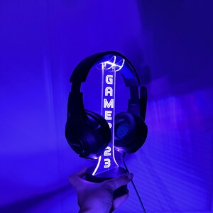 May include: A black gaming headset with a white LED stand that says "GAME 23". The stand is lit up in a bright blue light.