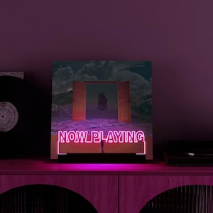 May include: A neon sign with the words "NOW PLAYING" in pink illuminates a vinyl record album cover. The album art features an open door leading to a surreal landscape. A vinyl record and a record player are visible.