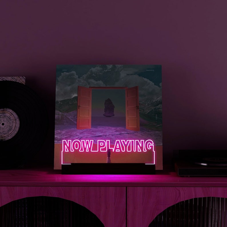May include: A neon sign with the words "NOW PLAYING" in pink illuminates a vinyl record album cover. The album art features an open door leading to a surreal landscape. A vinyl record and a record player are visible.