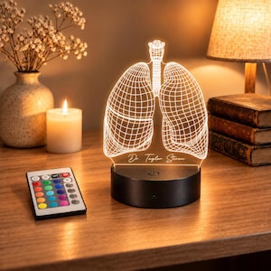Op de afbeelding: Een 3D LED-lamp met een witte draadmodel-illustratie van menselijke longen op een helder acrylpaneel. De voet is zwart en cilindervormig met een aan-uitknop. De tekst Dr. Taylor Steven is erin gegraveerd. Inclusief een afstandsbediening.