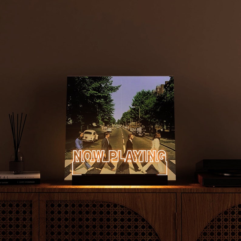 May include: Illuminated sign featuring the iconic Abbey Road album cover with the text "NOW PLAYING" in white. The image shows the Beatles crossing the street. The sign is on a wooden cabinet.