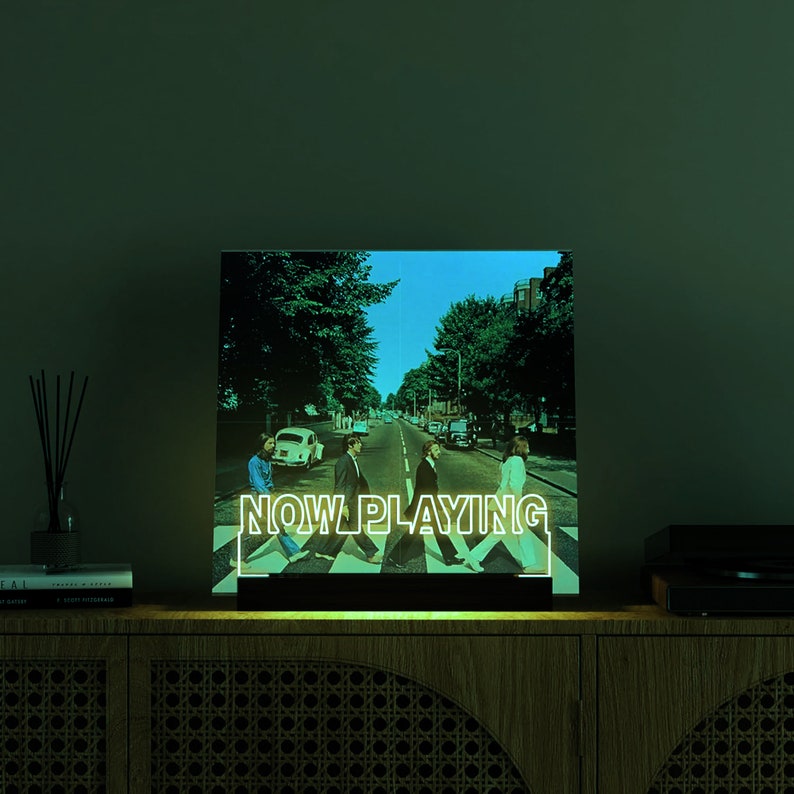 May include: Illuminated sign featuring the iconic Abbey Road album cover with the text "NOW PLAYING" in white. The image shows the Beatles crossing the street. The sign is on a wooden cabinet.