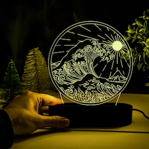 Puede incluir: Un diseño de arte de línea en blanco y negro de una ola rompiendo contra una costa con un sol brillando por encima. El diseño está iluminado desde atrás, creando un efecto brillante. El diseño está sobre una base negra.