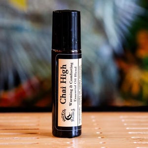 May include: A black essential oil roller bottle with a label that reads "Chai High" and "Warming & Comforting Essential Oil Blend." The bottle is on a wooden surface with a blurred background of green, orange, and blue.