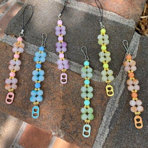 May include: Six colorful phone charms with a double ring design. Each charm features a gradient of pastel colors, including pink, blue, green, and yellow. The charms are made with small round beads and have a gold-colored ring at the bottom.