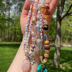 May include: Multi-strand beaded necklace with charms and letter beads spelling out "SASS DETECTIVE TELLIG" and "DEPUTY YOUR" in white letters on a white background.