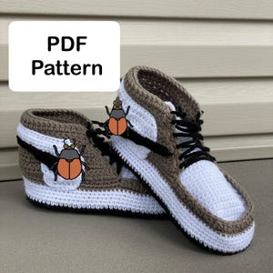 Puede incluir: Un par de zapatillas de deporte de ganchillo en marrón, blanco y negro. Las zapatillas tienen un diseño de caña alta con cordones negros y una suela blanca. El texto "PDF Pattern" se muestra en un rectángulo blanco. También hay escarabajos de dibujos animados decorativos.