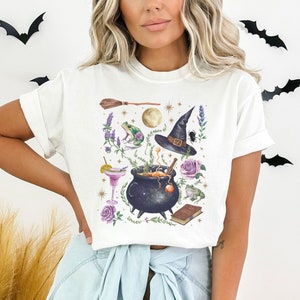 May include: White t-shirt with a graphic print of a witch's cauldron, a witch's hat, a frog, a broom, and other magical elements.