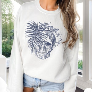 May include: White sweatshirt with a blue graphic of a skull with a palm leaf and butterflies. The skull is partially obscured by the leaf.