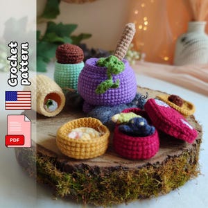 May include: A selection of brightly coloured, crocheted miniature food items, including a purple pot with a stem, a yellow bowl, and a red container. The items are displayed on a wooden surface covered in moss. The image includes the text "Crochet Pattern" and a PDF icon.