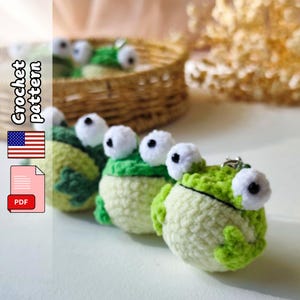 May include: Crocheted frog keychains in a row, featuring white and black eyes, green and light green bodies. The keychains are on a white surface, with a woven basket in the background. The image also includes a "Crochet Pattern" graphic with a US flag and a PDF icon.