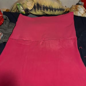 May include: A bright pink, folded piece of fabric, possibly a skirt or a similar garment, is the primary focus. The fabric appears to be a solid color, with some lighter areas. The background includes a large fish-shaped pillow and other items.