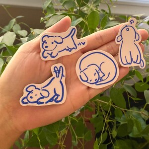 以下が含まれることがあります： 白いステッカーに青い線で描かれた犬のイラストが4つ。犬は走ったり、寝たり、座ったりと様々なポーズをとっています。