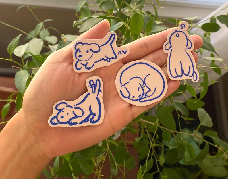 以下が含まれることがあります： 白いステッカーに青い線で描かれた犬のイラストが4つ。犬は走ったり、寝たり、座ったりと様々なポーズをとっています。